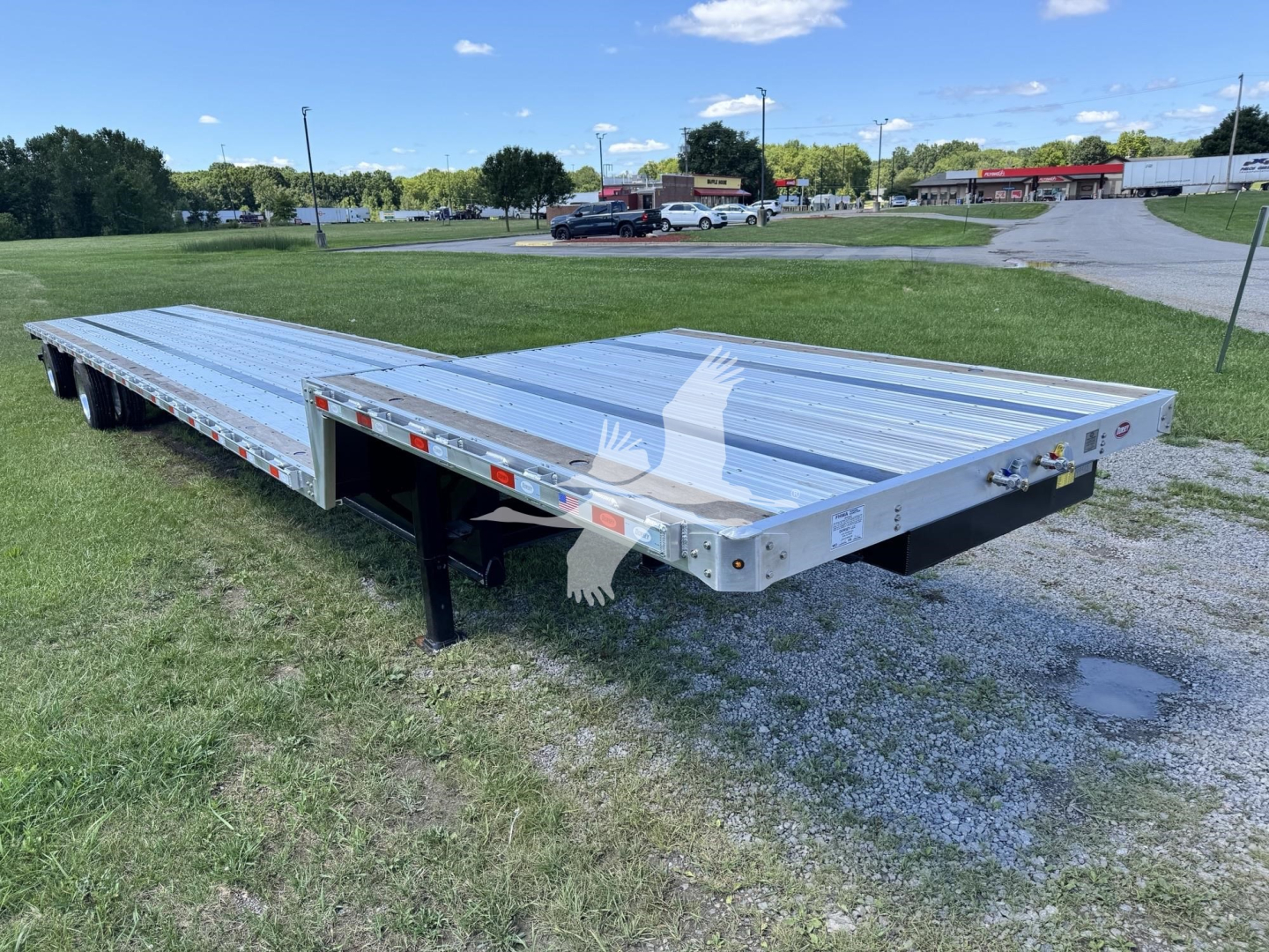 Flatbed trailer with a long aluminum deck and dual drop axles, viewed from the side on a grassy lot; appears new with reflective safety markings along the sides.