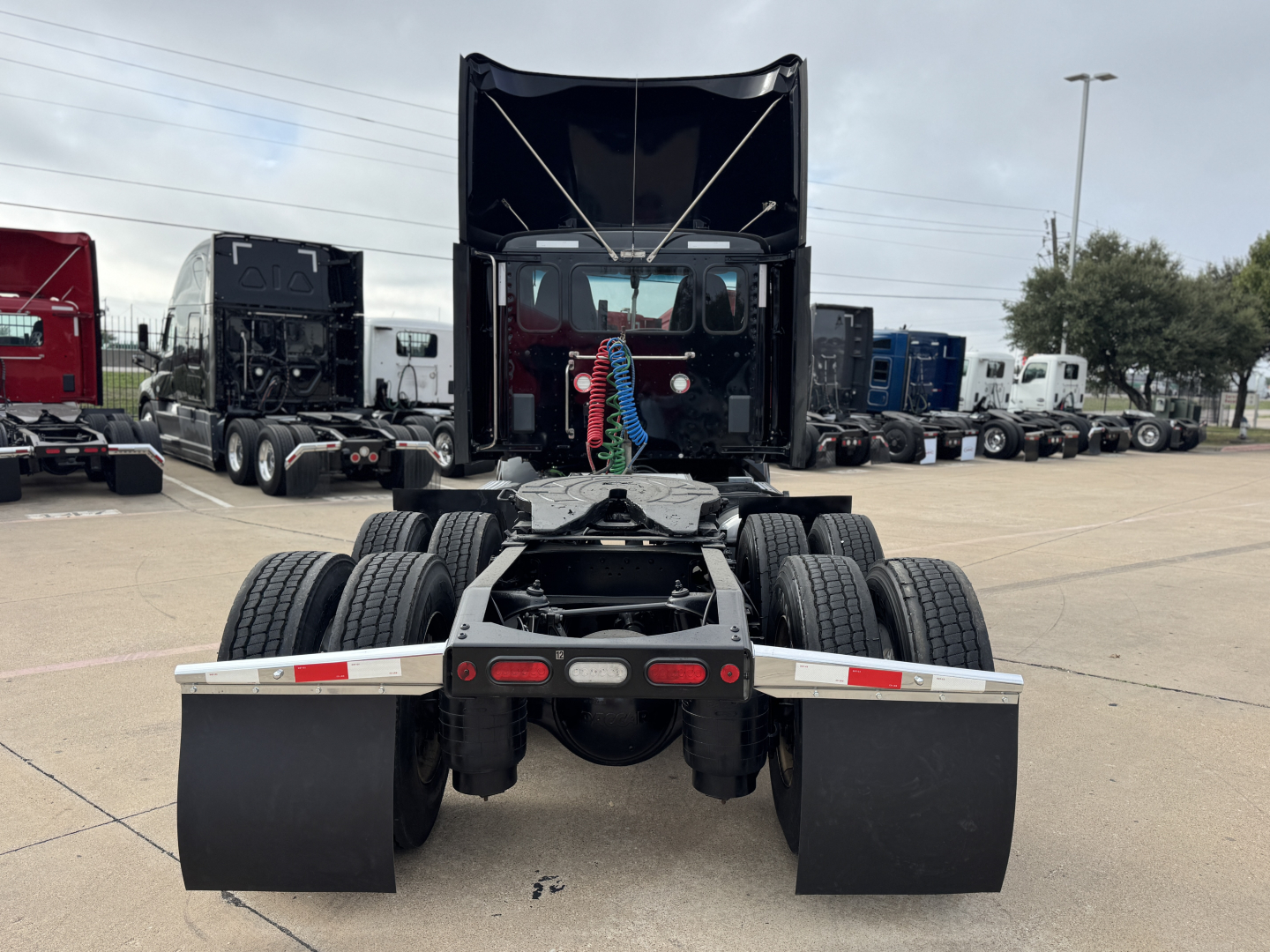 Peterbilt cab and chassis rear view showing dual rear axles and mudflaps with fifth wheel area; show truck bed and stacked trucks in background.