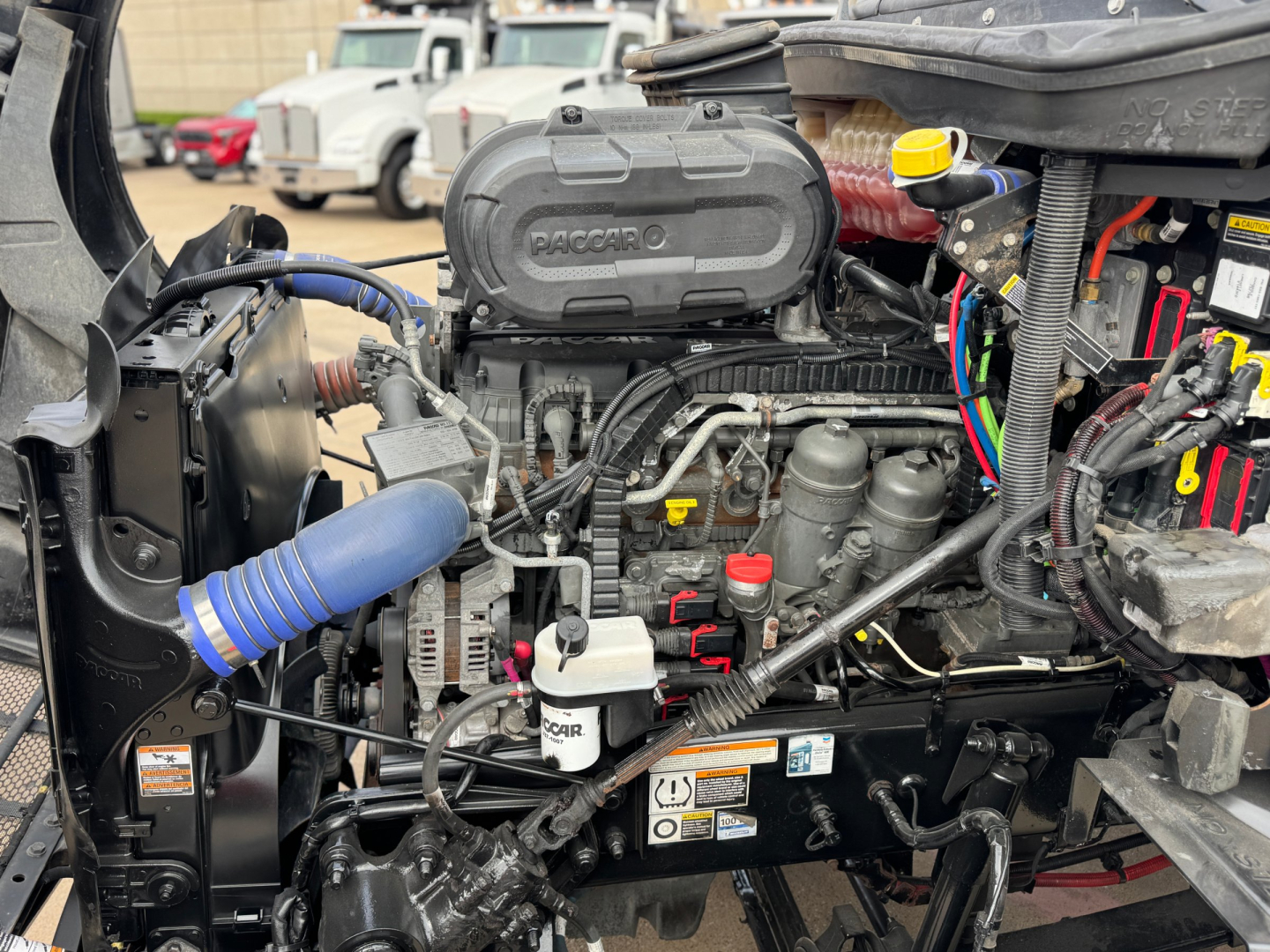 Peterbilt engine bay showing 6-cylinder engine with black intake cover labeled 'PACCAR' and various hoses, wiring, and reservoirs visible.