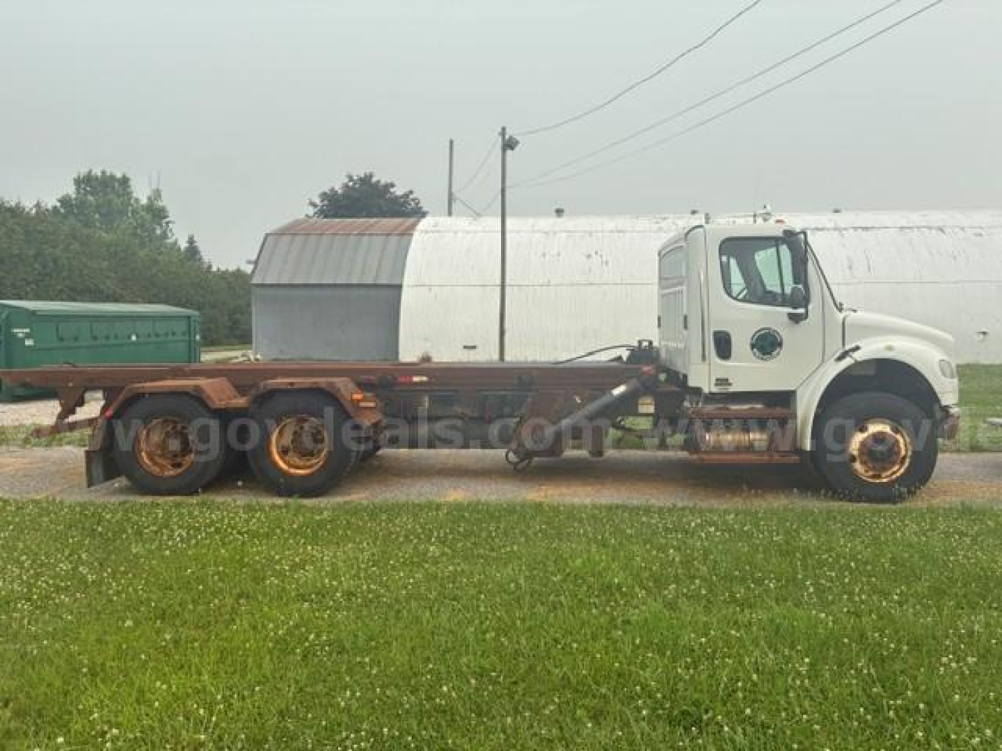 Cab and chassis truck in profile showing Freightliner M2 106 chassis with white cab and long flatbed, cargo trailer in background, license-free rural lot.