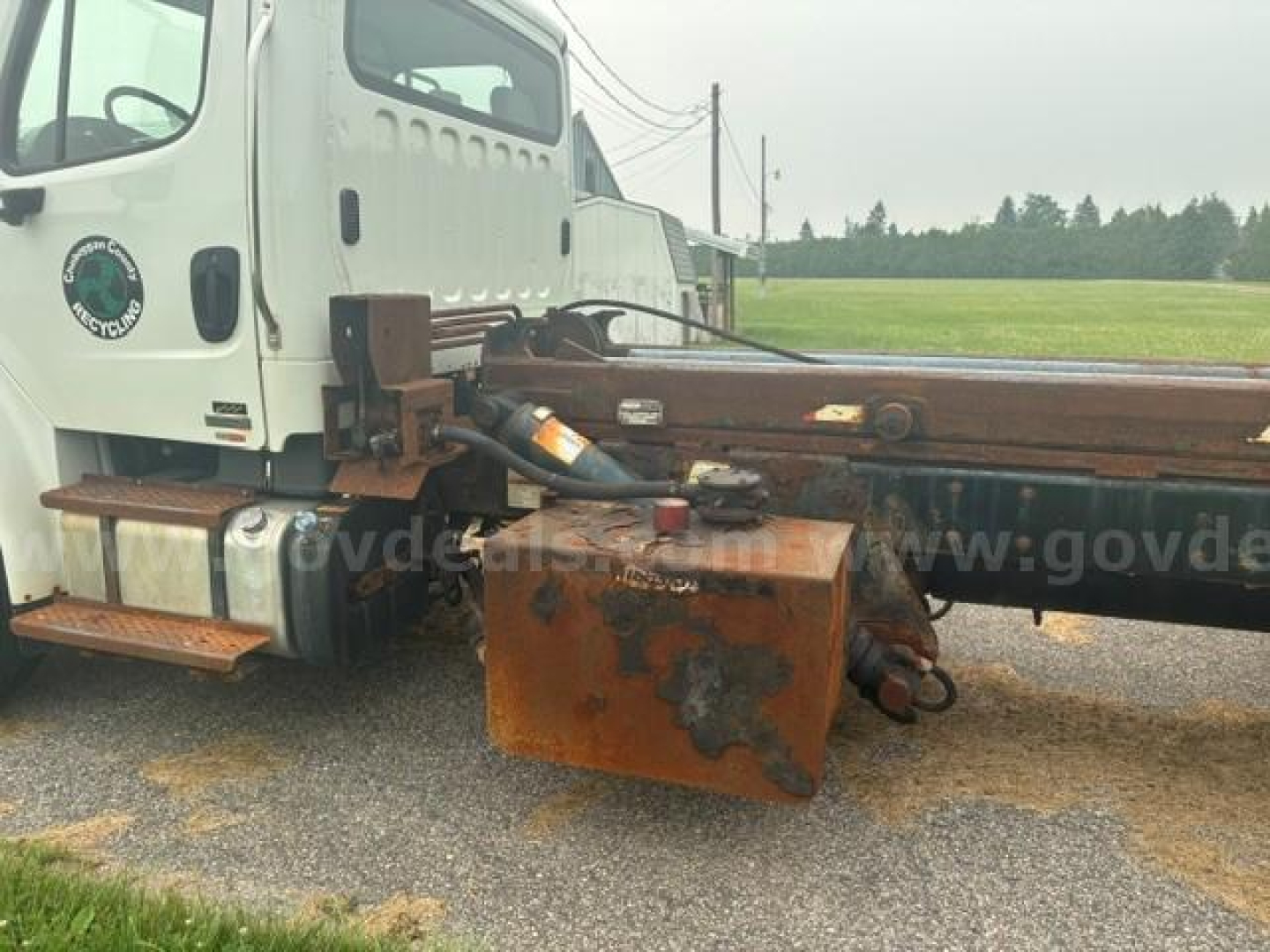 Truck cab and chassis of a 2012 Freightliner M2 106 with rusty equipment under the flatbed rail, side view showing fuel tank and mounting.
