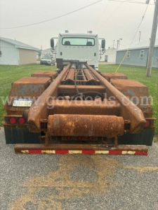 Rear view of a Freightliner M2 106 cab and chassis with a rusty hook-lift trailer setup and long frame extending toward the cab.