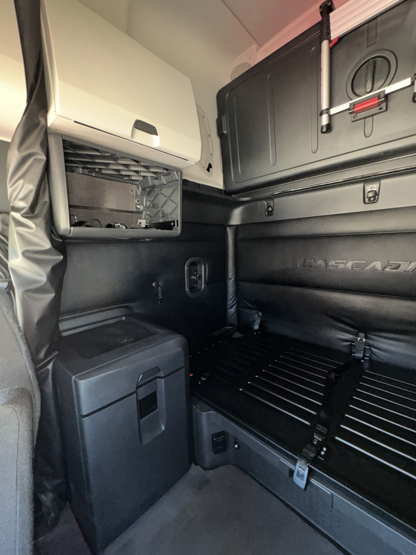 Cab interior of a Freightliner Cascadia 126 sleeper showing storage compartments, side panel, and safety rails; appears to be a used truck with black interior trim.