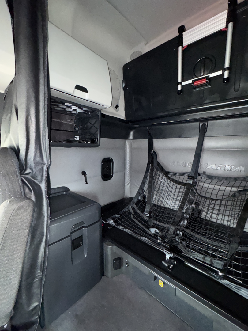 Truck sleeper cab interior showing bunk area with cargo net, storage cabinet, and seating; compact layout and fixtures visible.