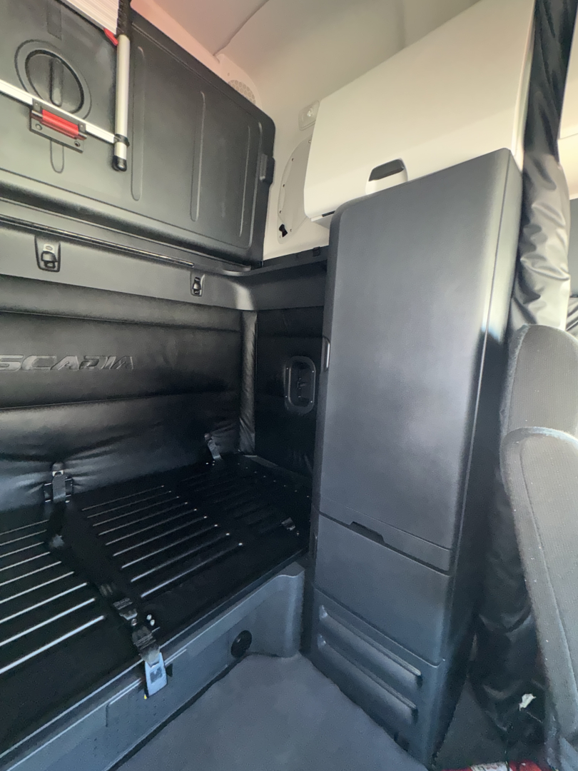 Freightliner Cascadia sleeper cab interior showing storage compartments and partial rear seating area with black panels and overhead storage.