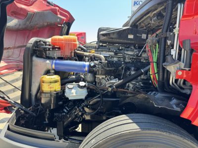 Truck engine bay showing coolant reservoir with orange cap, blue silicone hose, yellow filter, hydraulic lines, and exposed engine components.