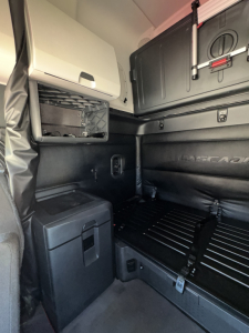 Cab interior of a Freightliner Cascadia 126 sleeper showing storage compartments, side panel, and safety rails; appears to be a used truck with black interior trim.