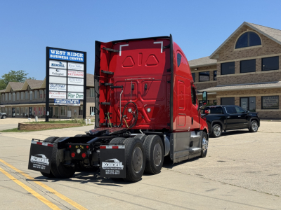 Truck chassis and red sleeper cab of a 2023 Freightliner Cascadia 126 from rear-left angle, with twin rear axles and mud flaps visible.