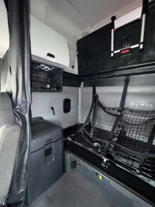 Truck sleeper cab interior showing bunk area with cargo net, storage cabinet, and seating; compact layout and fixtures visible.