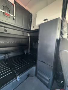 Freightliner Cascadia sleeper cab interior showing storage compartments and partial rear seating area with black panels and overhead storage.