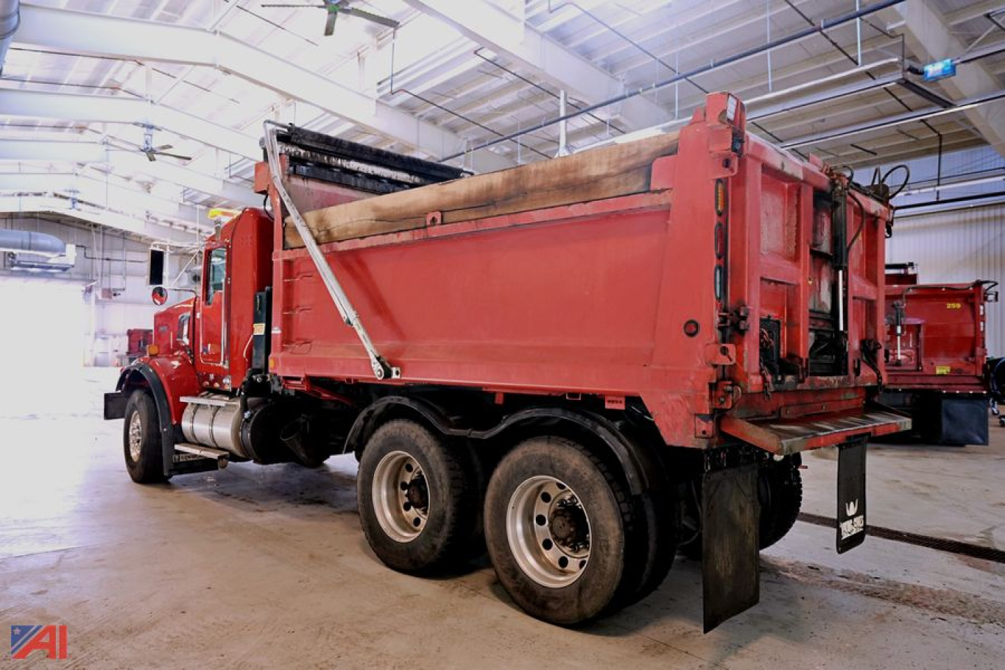Kenworth T8 Series dump truck in a warehouse, red body, rear dump bed, dual rear axles, interior lighting visible.