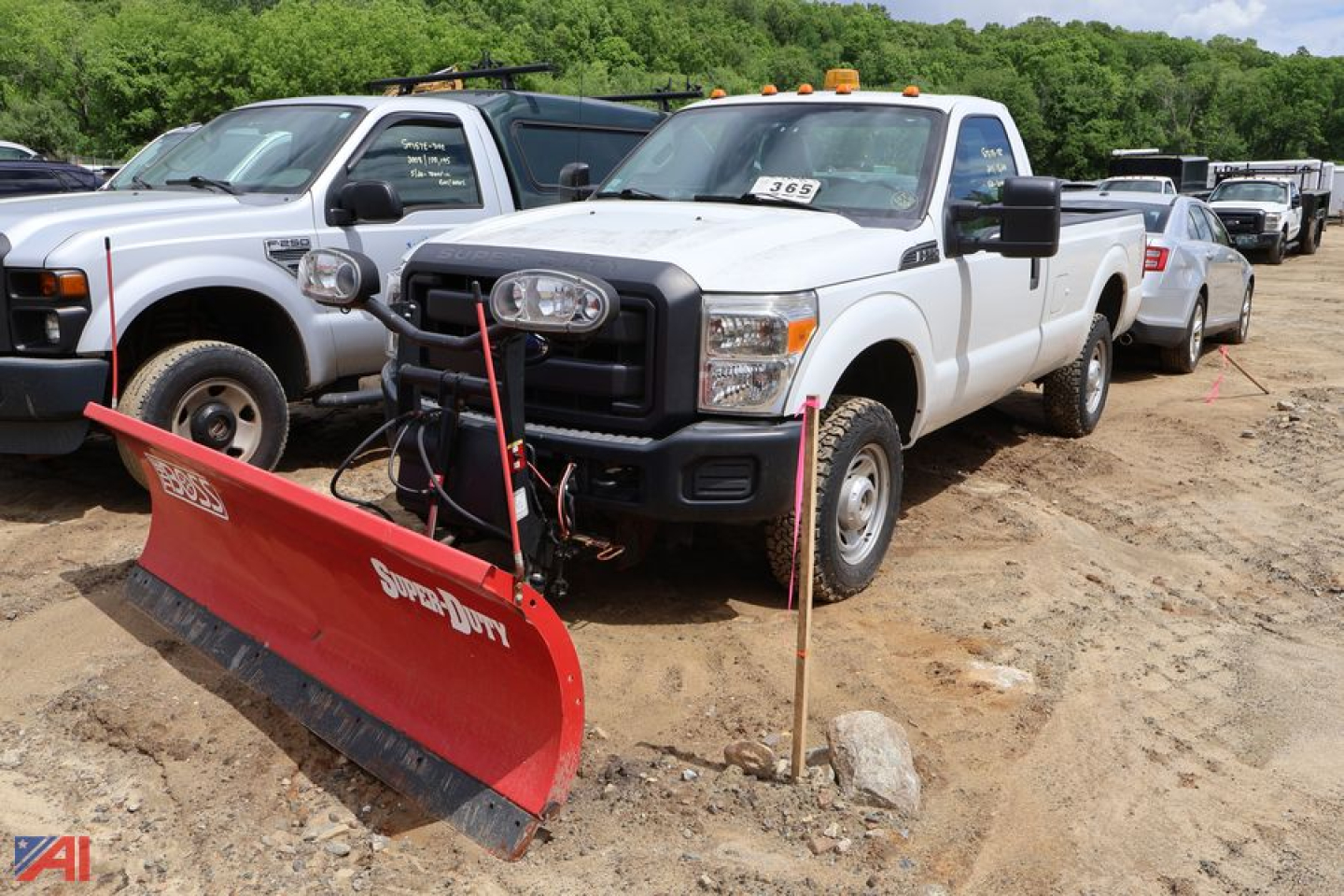 Front view of a white Ford F-250 Super Duty pickup with a red snow plow attached, other trucks in background, on dirt lot.