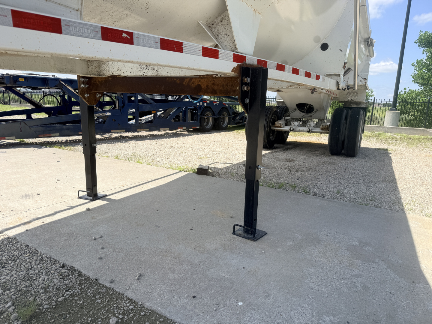 Trailer underside with ground supports extended, showing legs and side rail of a dump trailer in service; text visible on side rails