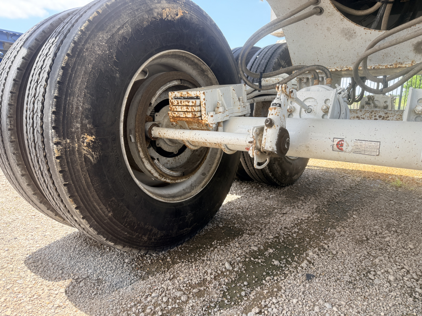 Truck trailer axle showing dual rear wheels and suspension components with rust on brake chamber area and side view of axle beam.