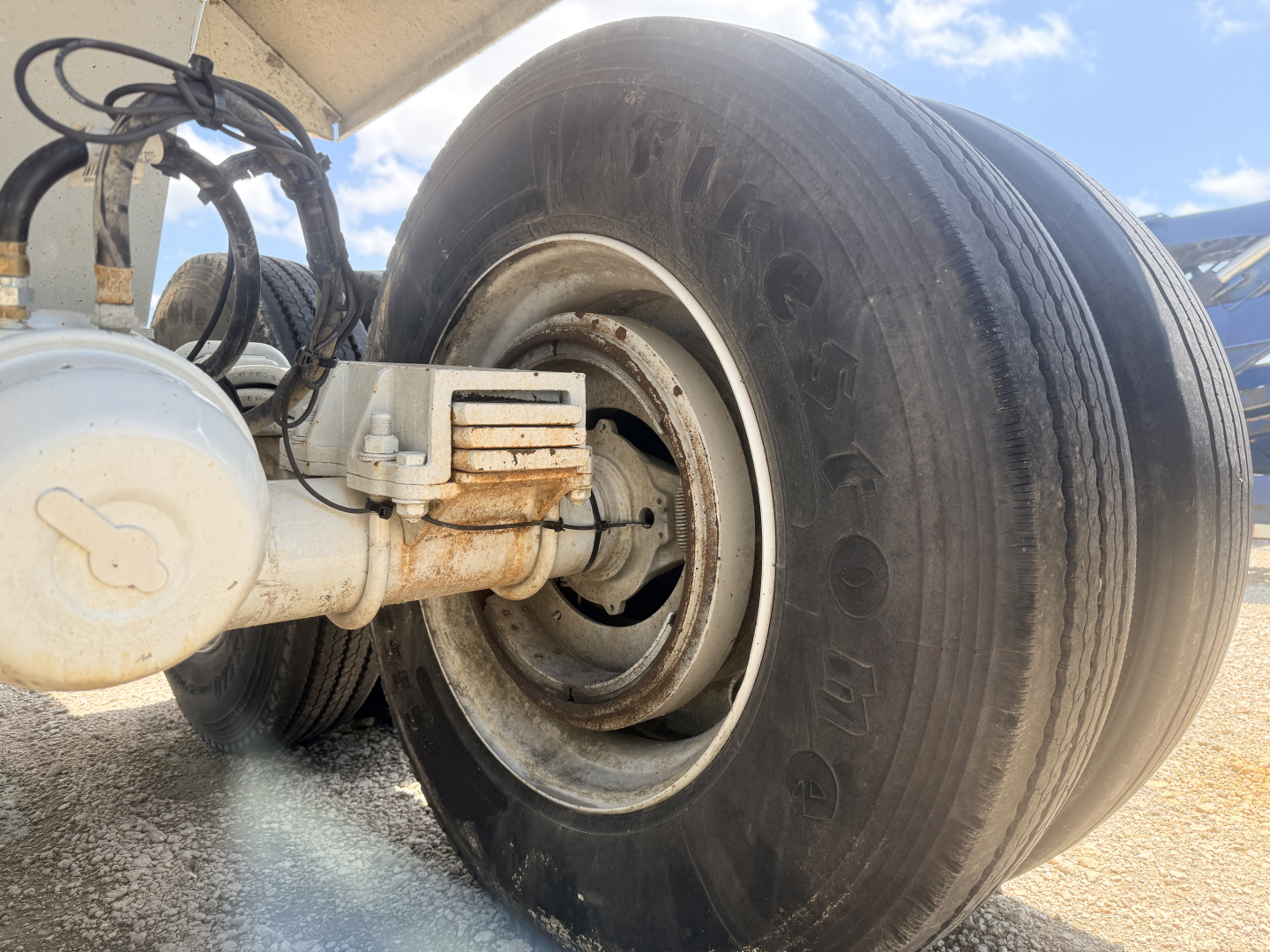 Truck trailer axle showing dual tires and suspension with air brake chamber and leaf spring. Visible tire sidewall reads 'Goodyear'.