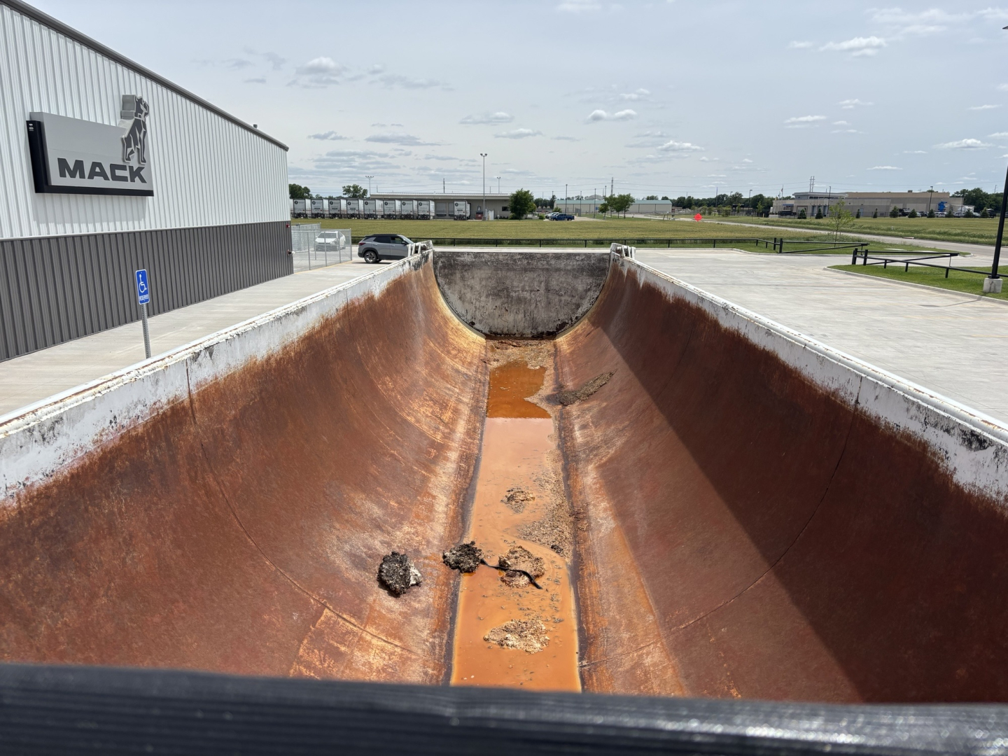 Dump trailer bed interior showing rusted metal walls and rusty floor with debris, Mack dealership building in background