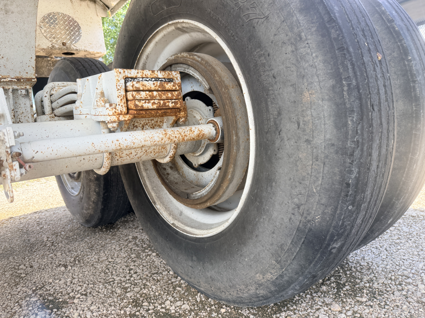 Front axle and wheel assembly of a dump trailer showing alloy rim, large tire, and rusty hitch mechanism.