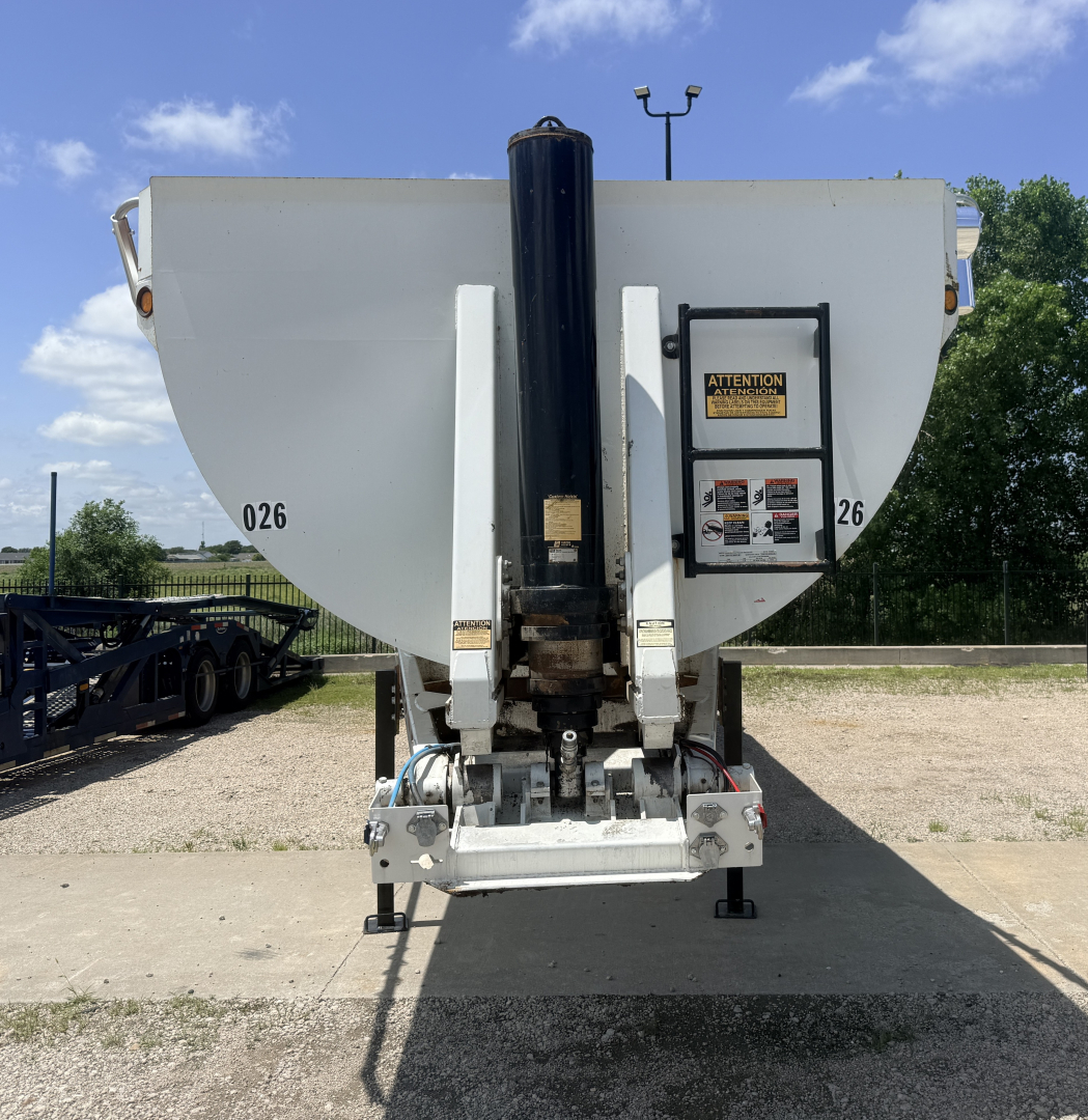 Dump trailer front view showing hydraulic cylinder and hitch with warning label, gray body, side numbers 026, ID plate and support legs.