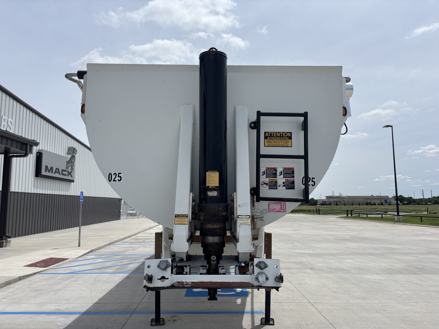 Dump trailer hitch with hydraulic lift mechanism and open-top bed. Visible labels 025 on sides, black hydraulic column center, warning label panel on right side.