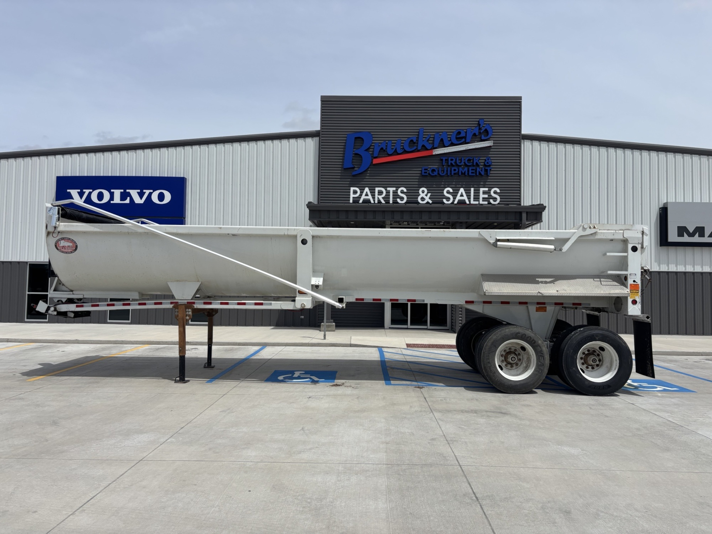 Dump trailer with long white body and dual rear axles parked in front of Braackner truck equipment building showing Volvo sign in background.