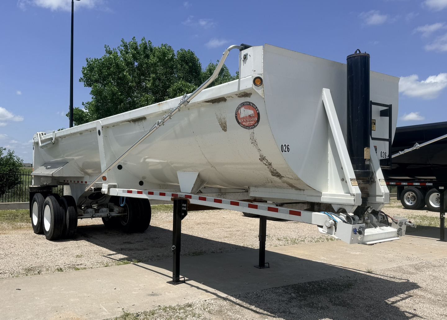 Dump trailer showing side view with white steel body, dual rear axles, landing legs, and hitch with safety chains.