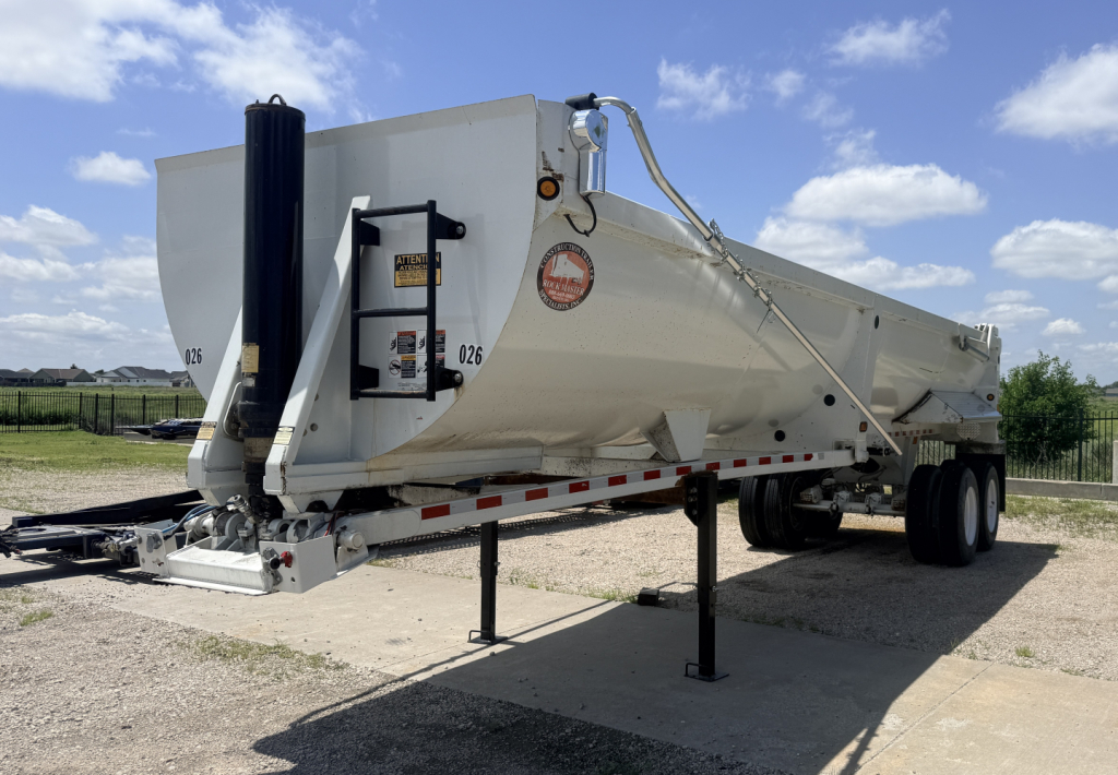 Dump trailer showing side view with white body, tandem axles, front gooseneck hitch, and stabilizing leg extended.