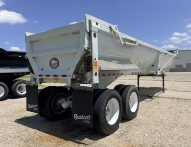Dump trailer with dual tandem axles and white sides, rear lights, and Bruckners mudflaps showing spare wheel, resting on gravel.