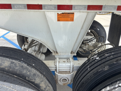 Dump trailer undercarriage showing tandem axles, suspension, and hitch area. Visible text on orange sticker and reflective red-white tape along top edge.