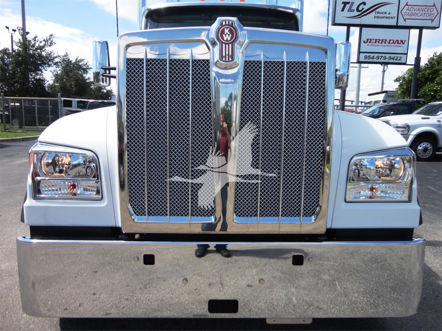 Front view of a 2022 Kenworth W900 truck with chrome grille, large bumper, and emblem, driving a rollback truck lot.