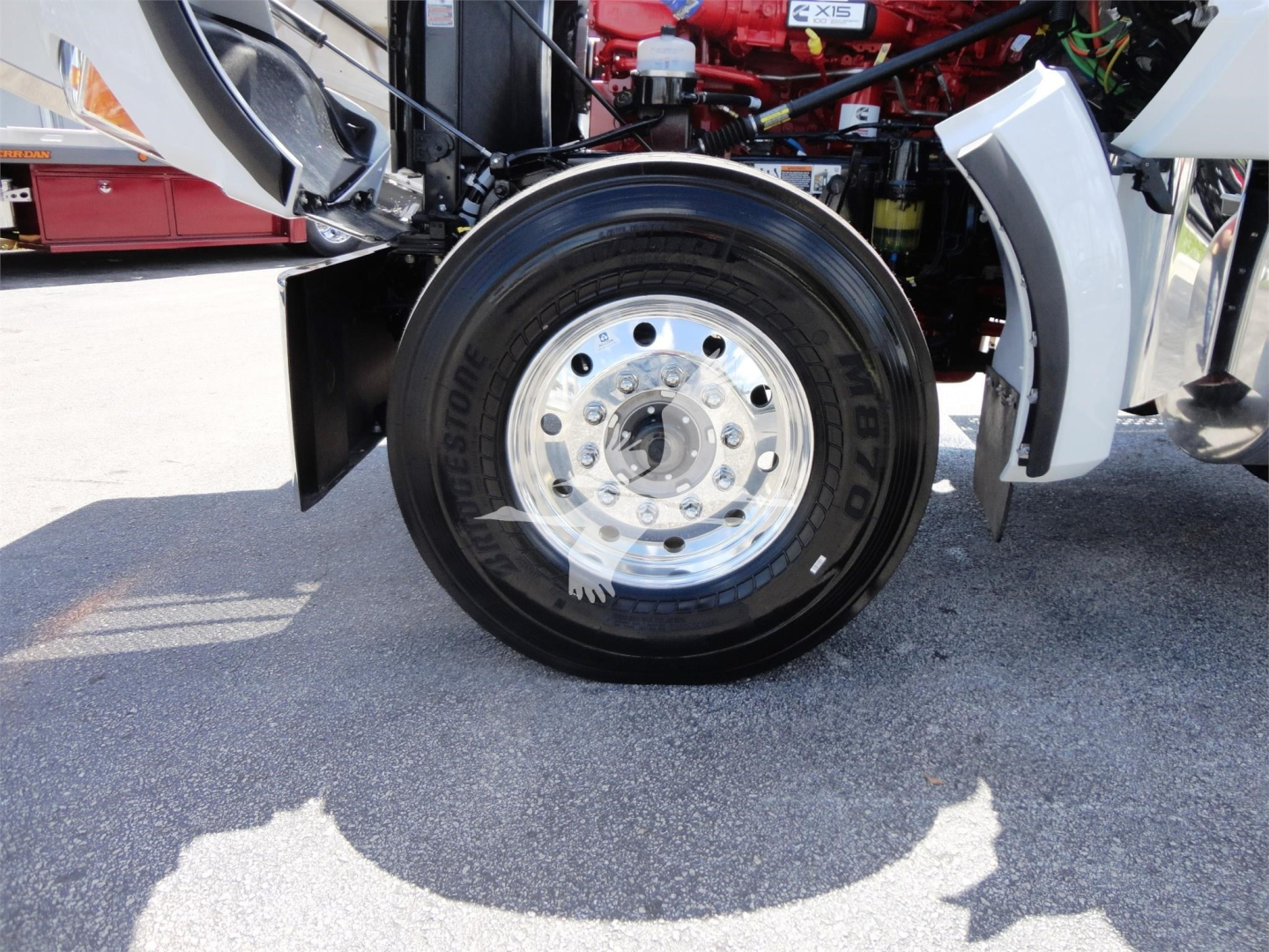 Kenworth W900 front wheel with polished aluminum hub, black Goodyear tire, and exposed red engine components in the background.