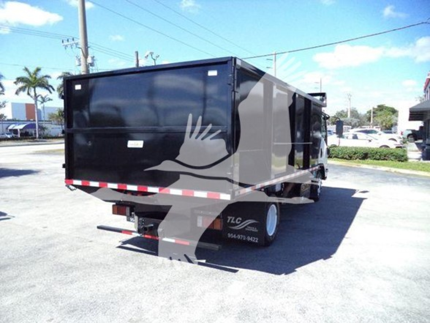 Isuzu NPR dump truck trailer showing black dump body with white and red reflective striping and TLC wheel cover in a parking lot.