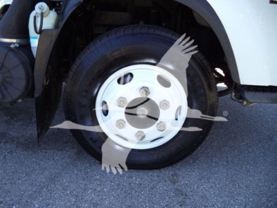 Isuzu NPR wheel and front fender area showing tire and white hubcap with lug nuts; visible component includes part of wheel well and a coolant reservoir behind.