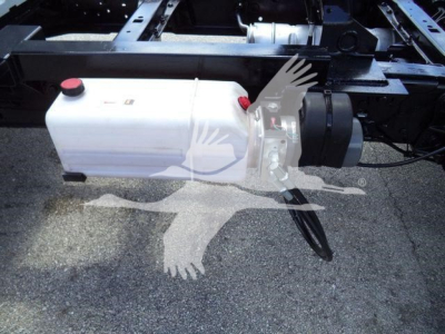 White coolant reservoir with red cap mounted on truck frame, showing plumbing and mounting hardware; portion of chassis and hoses visible.