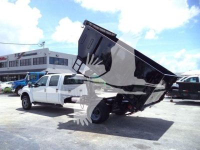 Ford F-550 dump truck with raised dump bed in a parking lot, white cab, black bed, blue sky, dealership in background.
