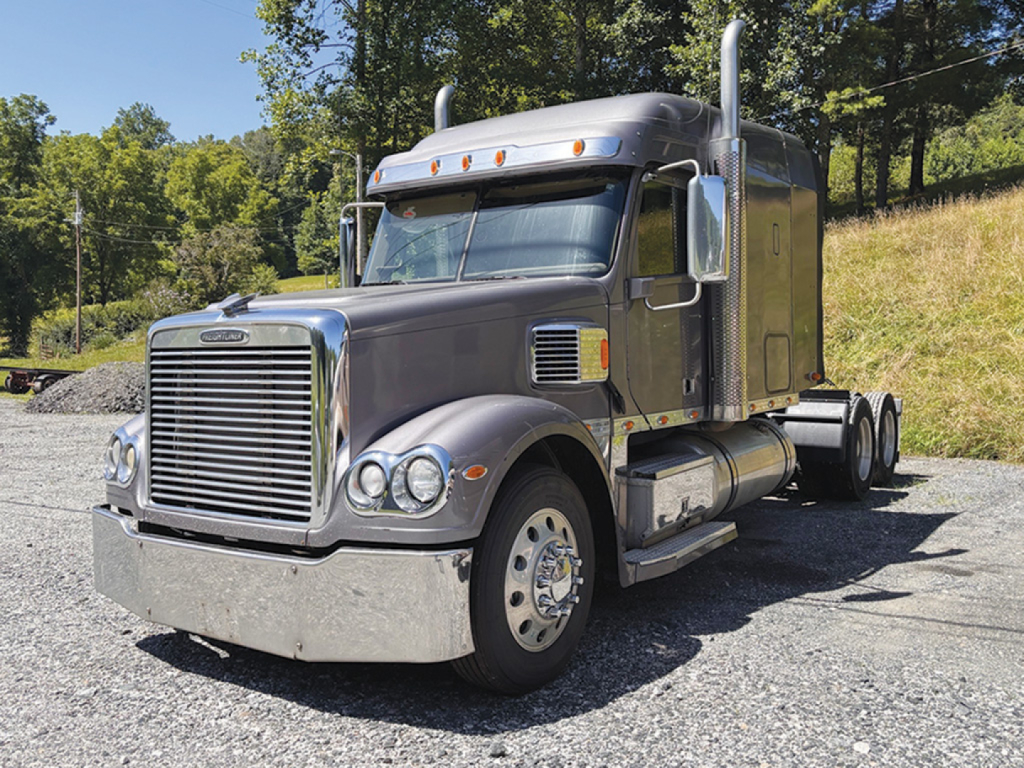 Truck cab and front end of a 2015 Freightliner Coronado conventional sleeper tractor, metallic gray with chrome grille, dual exhaust stacks, and polished fuel tanks.