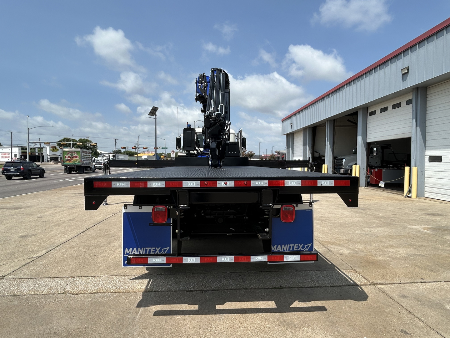 Rear view of a new Mack GR64F crane truck with a mounted crane, Manitek blue mud flaps, red and white reflective stripes, and a service bay in background.