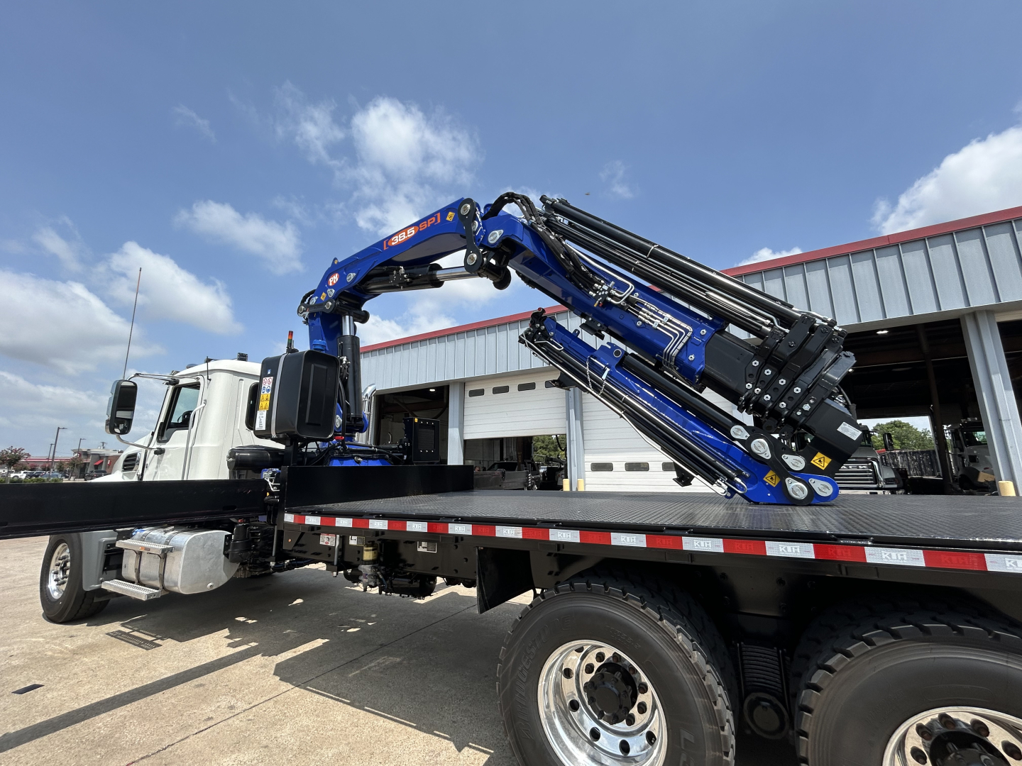 Truck crane in service bay showing hydraulic knuckle-boom crane mounted on a flatbed truck with Mack GR64F nameplate and blue arm components.
