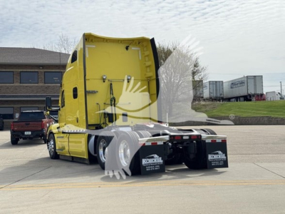 Peterbilt 579 tractor back view showing faded yellow sleeper cab and rear wheels, trailer hitch, mud flaps with Konzel signs, parking lot, other trucks in background.