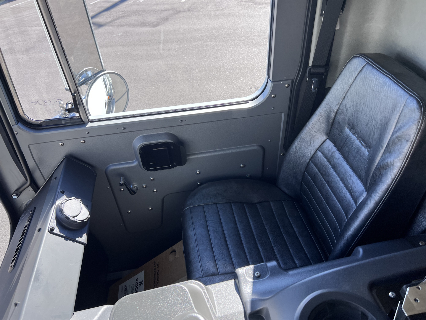 Cab interior of a Mack TE64 truck showing driver's seat, door panel with window, control knobs, and a small display panel; gray metal surroundings.