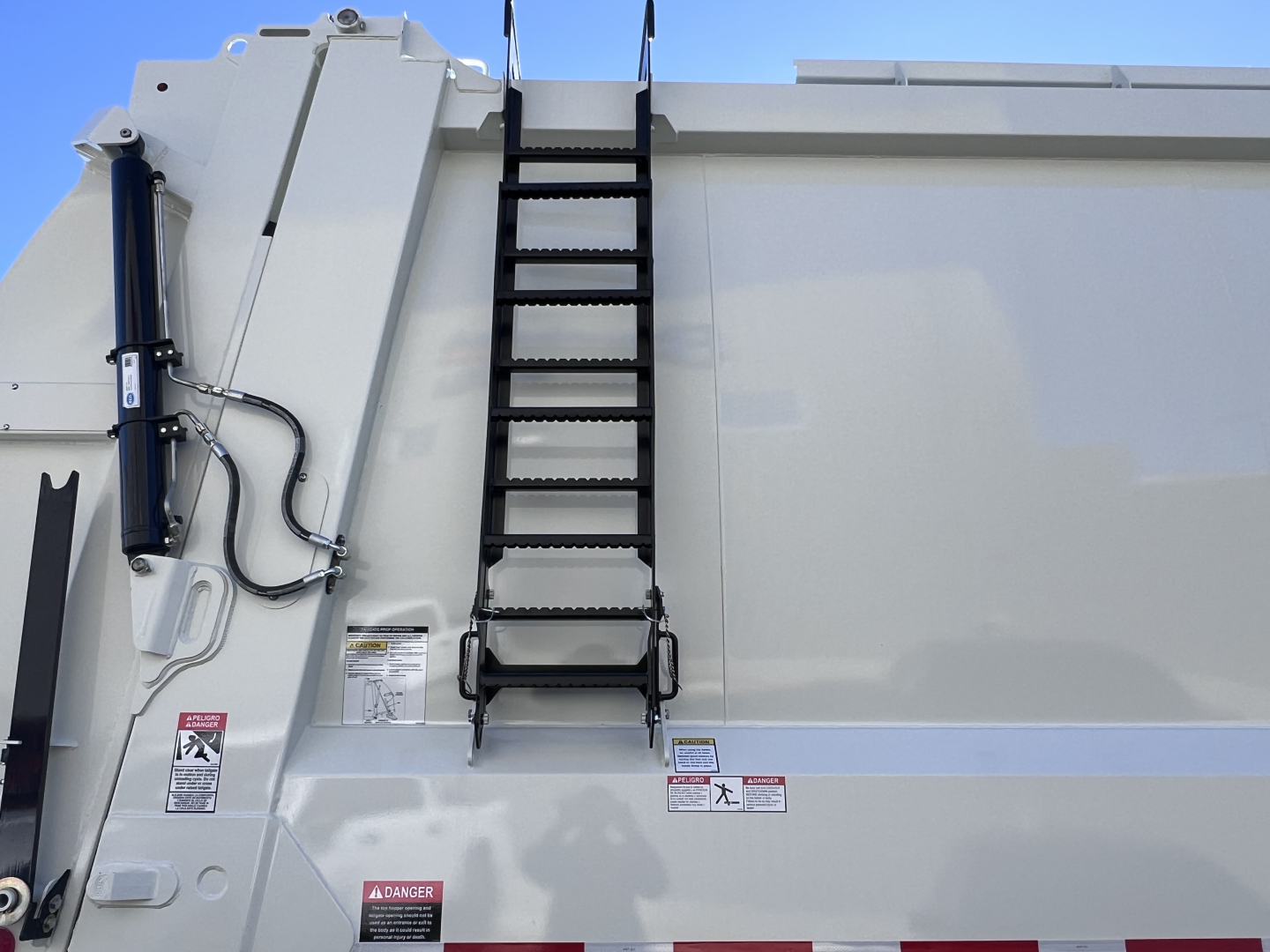 Truck side with vertical ladder on the dump body, hydraulic cylinder, and safety stickers visible.
