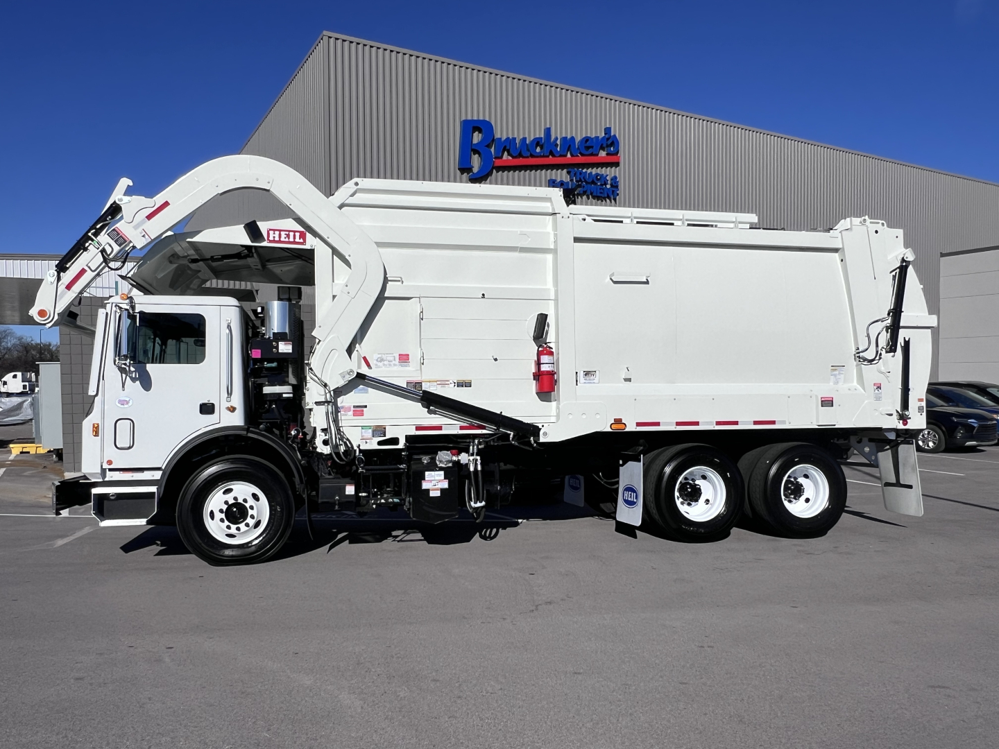 Mack TE64 refuse truck in service bay showing side view with dump body and hydraulic arm under bright blue sky.
