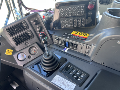 Cab interior of a Mack TE64 truck showing gear shifter, control panel, switches, and dashboard with diagnostic display and warning labels.