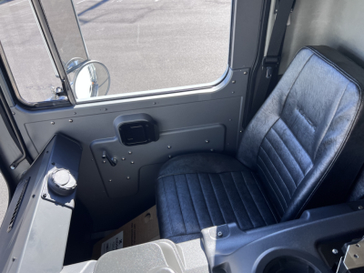 Cab interior of a Mack TE64 truck showing driver's seat, door panel with window, control knobs, and a small display panel; gray metal surroundings.