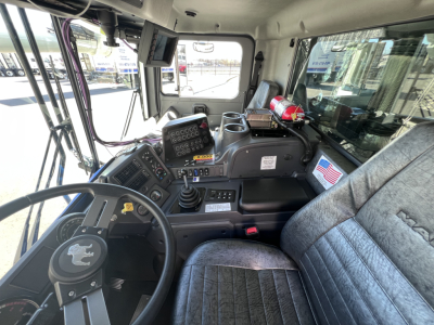 Mack TE64 cab interior showing steering wheel, instrument panel, gear shifter, multiple controls, monitor, and badge with U.S. flag.