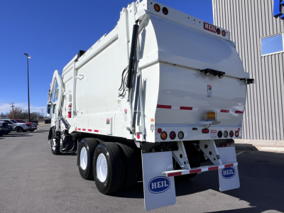 Rear and side view of a white Mack TE64 refuse truck showing dual rear axles, dump body, hydraulic lift, Heils mud flaps, and tail lights.