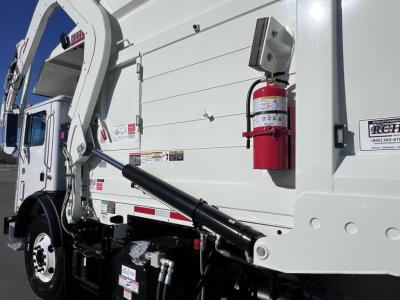 Trash truck side view showing hydraulic arm and white cargo body with red fire extinguisher mounted on side, safety decals, and black undercarriage.