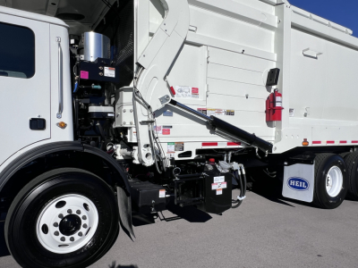 Truck front-left side of a 2026 Mack TE64 with hydraulic arm, dump body, and white cab; visible decals and label stickers on chassis.