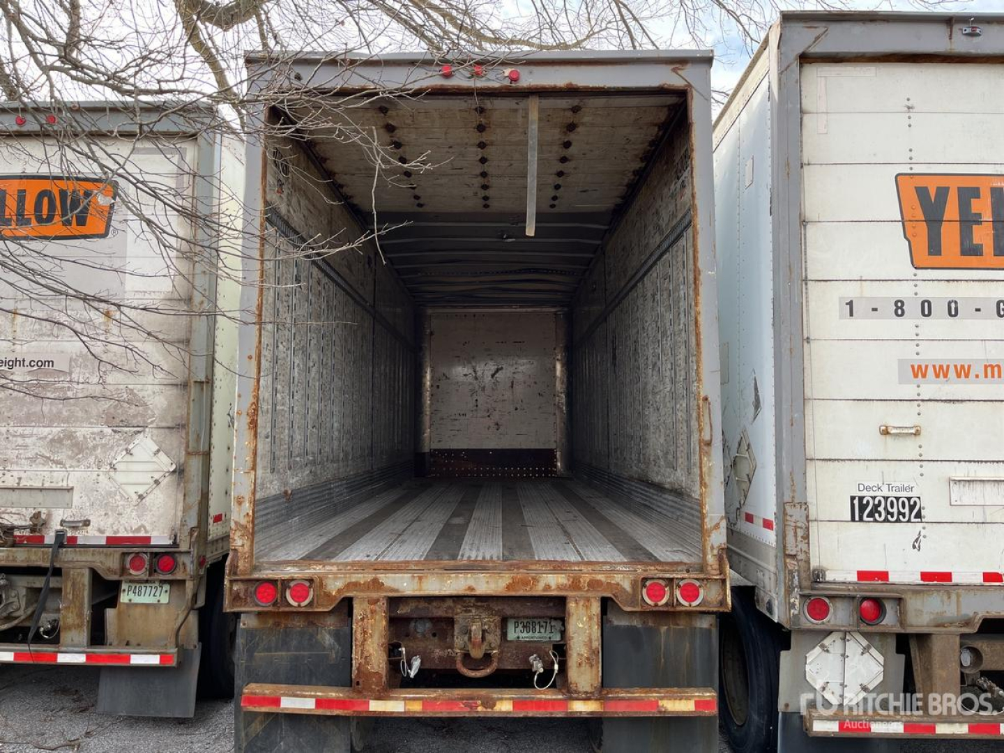 Trailer interior showing empty cargo space with metal floor and stripped walls; rear of trailer with tail lights and bumper visible; brand logos partially visible on adjacent trailers.