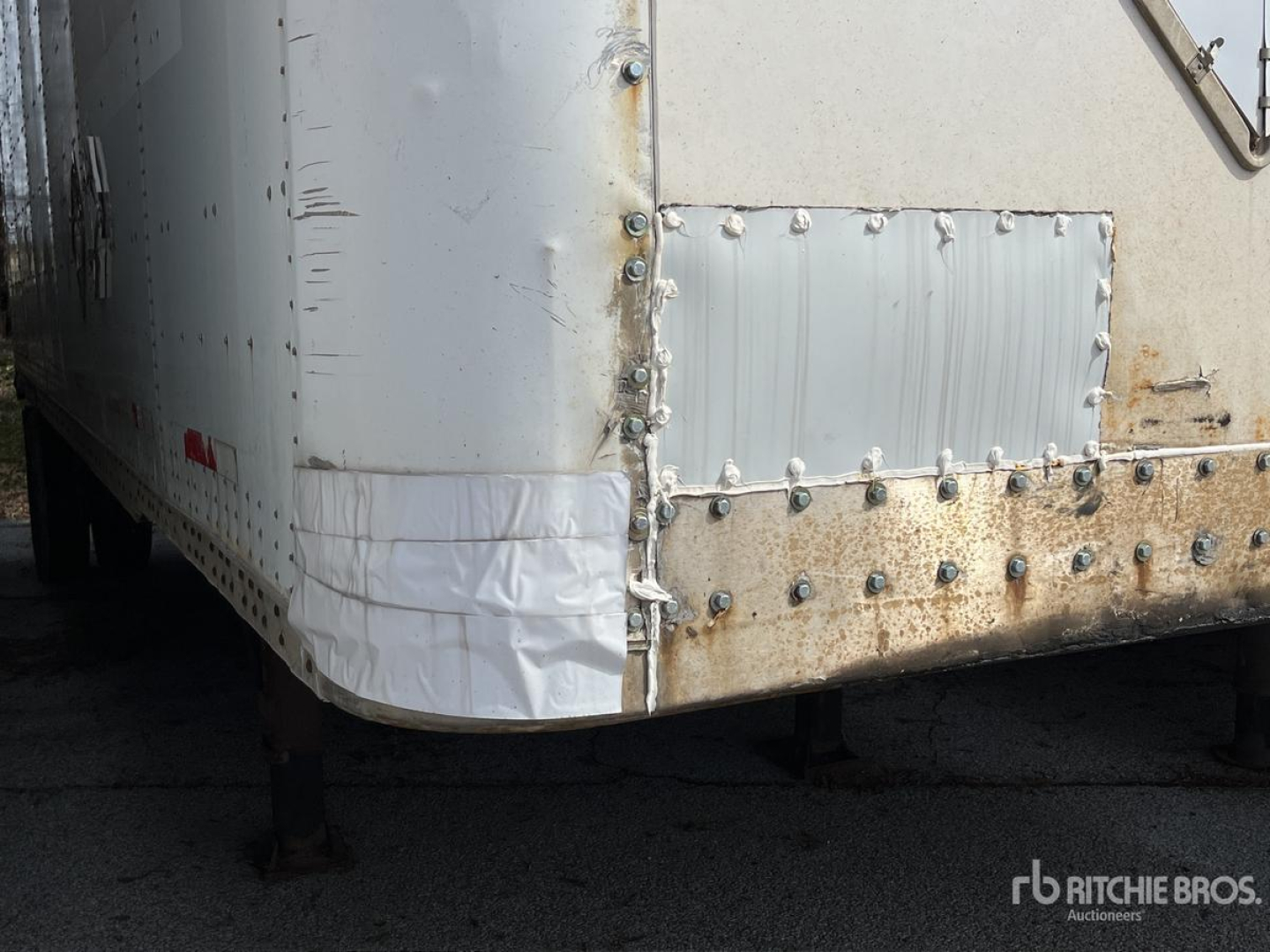 Trailer rear corner showing riveted metal panel with patch, wheel well edge, and foot support legs visible.