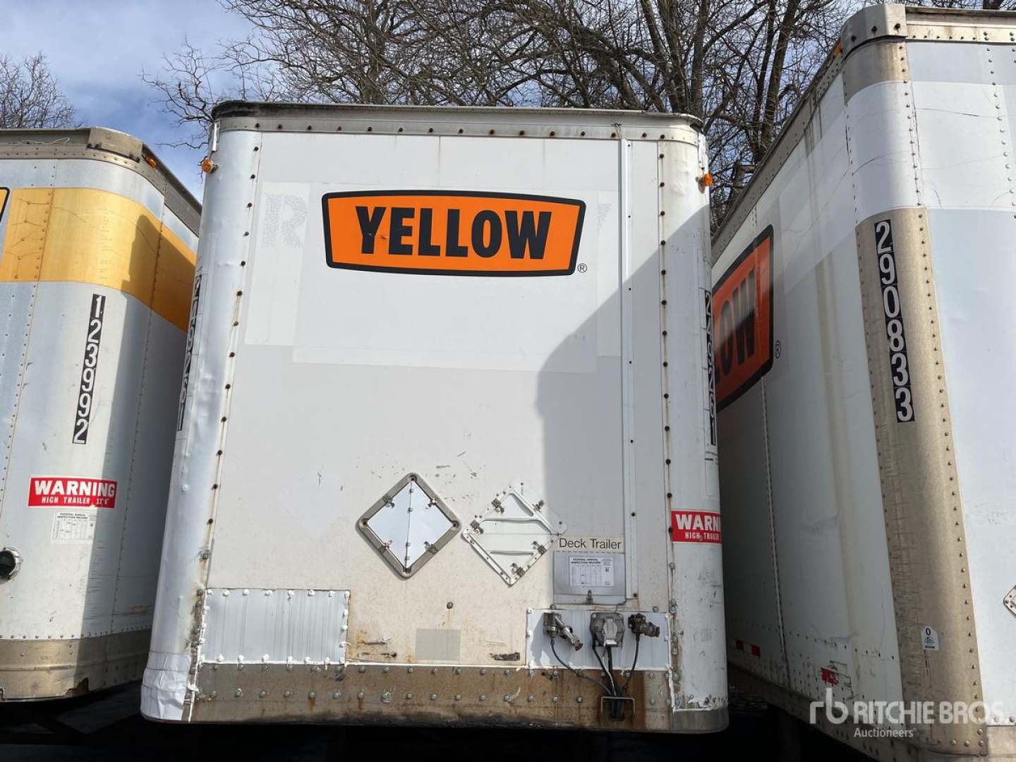 Rear of a white trailer with a yellow label reading 'YELLOW' mounted on the back doors; various safety stickers and metal hardware at the bottom; neighboring trailers on both sides.